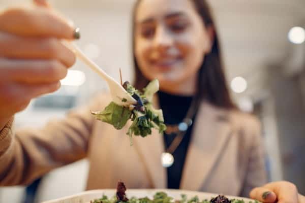 Girl Eats Vegetable Salad. Lady Sitting In A Cafe. Brunette In A Brown Coat